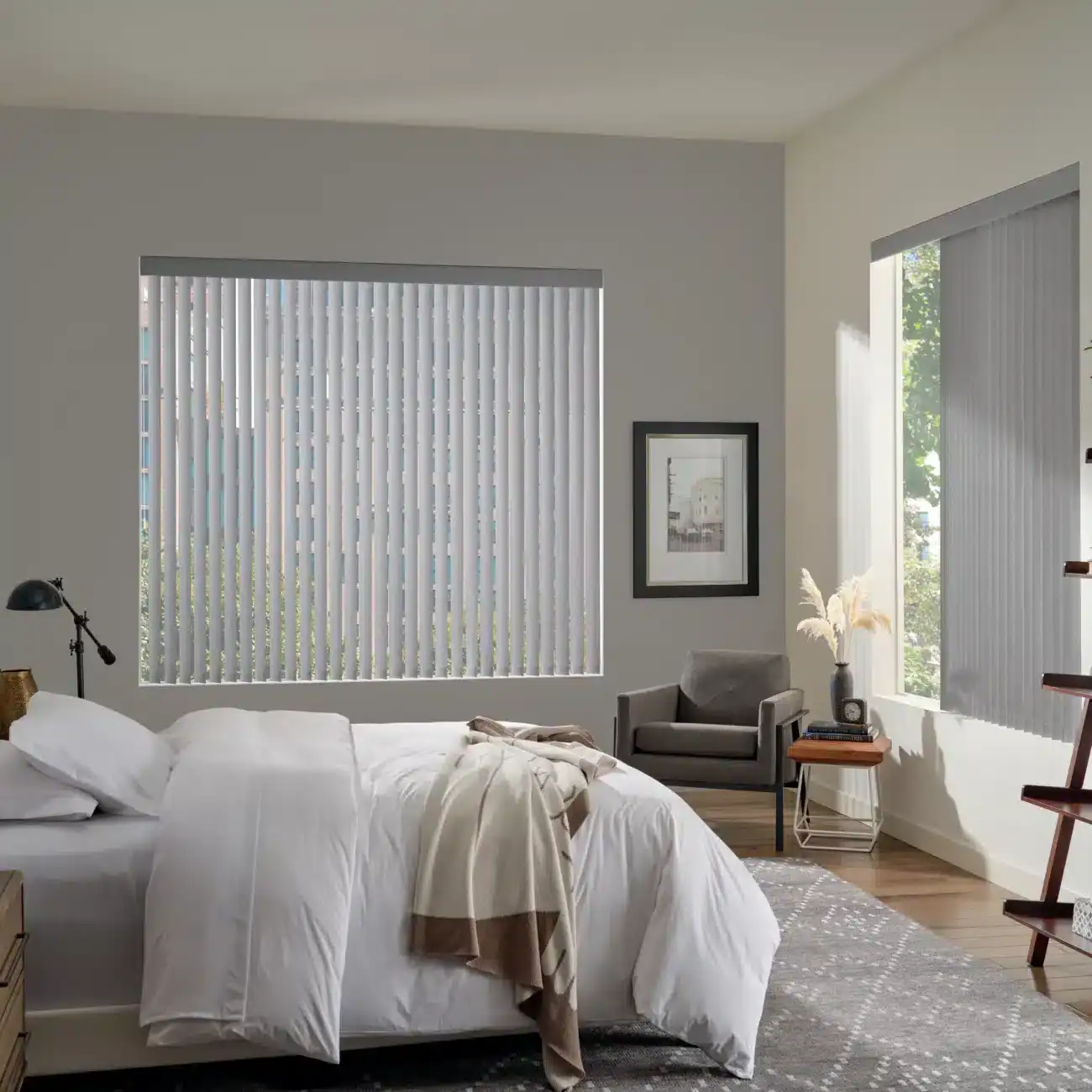 Modern bedroom with large windows featuring white vertical blinds, cozy bed, stylish furniture, and natural light, showcasing custom drapery solutions by Advance Blinds & Drapery.