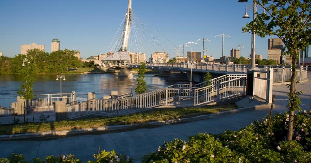 Bridge over river with modern architecture and city skyline in the background, featuring walkways, street lamps, and greenery, capturing urban waterfront scenery.