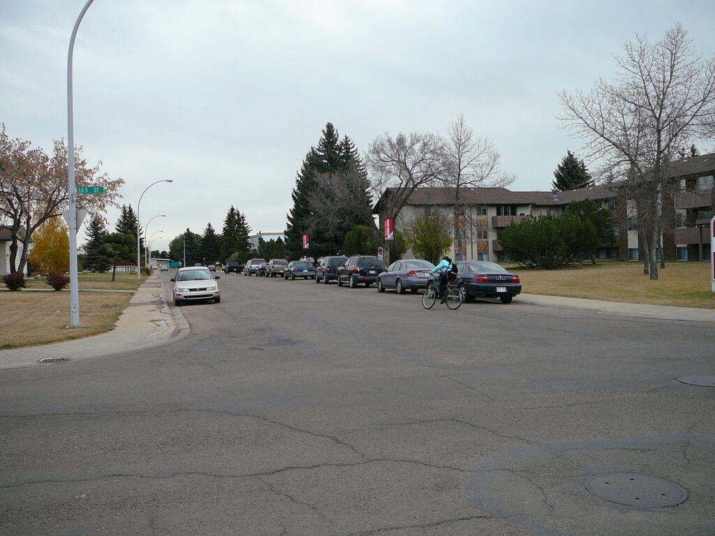 Driveway with parked cars leading to an apartment complex, featuring trees and a cyclist, with a focus on residential community and urban lifestyle.